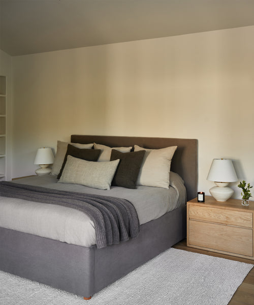 A modern bedroom featuring the Pacific Bed with a gray upholstered headboard, neutral bedding, and throw pillows. A wooden nightstand holds a lamp, candle, and flowers. Light walls and minimal decor create a soothing retreat.