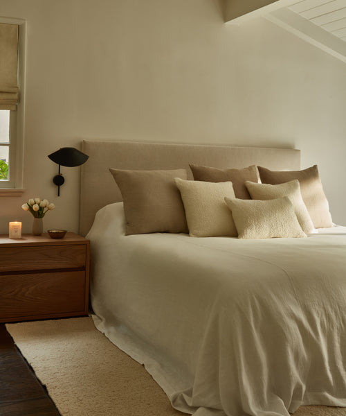 A cozy, neutral-toned bedroom featuring the Pacific Bed with a beige upholstered headboard, tan and cream pillows, a wooden nightstand holding a candle, tulip vase, small bowl, and soft natural light streaming through the window.