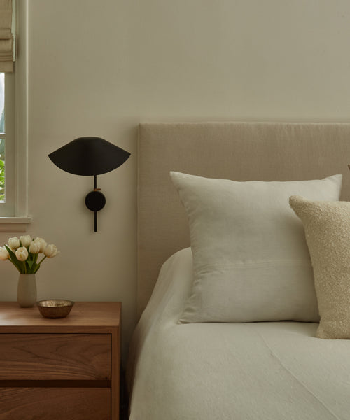 A minimal bedroom featuring a beige upholstered bed, white bedding, a Linen Pillow with a feather down insert, a wooden nightstand with tulips in a vase, a small dish, and a modern black wall sconce above the nightstand.