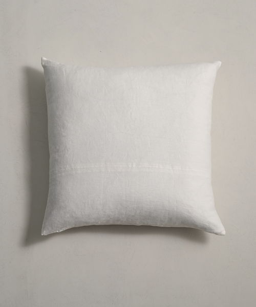 The Linen Pillow, featuring a feather down insert and plain white linen cover, is placed against a light grey textured wall and photographed from above.