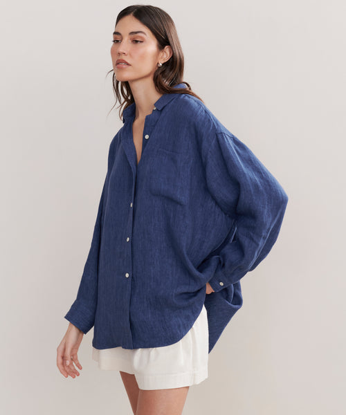 A woman wears the Relaxed Oversized Shirt, a loose-fitting, long-sleeve chambray top, with white shorts. She stands against a plain light background with one hand in her pocket, gazing off to the side.