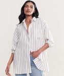 A woman wears the Linen Relaxed Oversized Shirt, showcasing vertical taupe stripes and relaxed button-up styling, paired with light blue jeans. She stands with one hand in her pocket against a plain, light background.