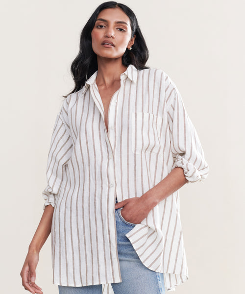 A woman wears the Linen Relaxed Oversized Shirt, showcasing vertical taupe stripes and relaxed button-up styling, paired with light blue jeans. She stands with one hand in her pocket against a plain, light background.