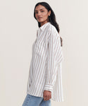 A woman models the Linen Relaxed Oversized Shirt with taupe stripes and blue jeans, standing sideways and looking at the camera against a plain light background.