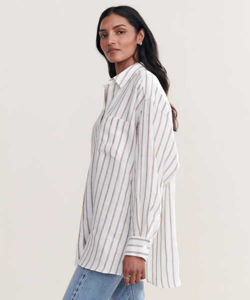 A woman models the Linen Relaxed Oversized Shirt with taupe stripes and blue jeans, standing sideways and looking at the camera against a plain light background.