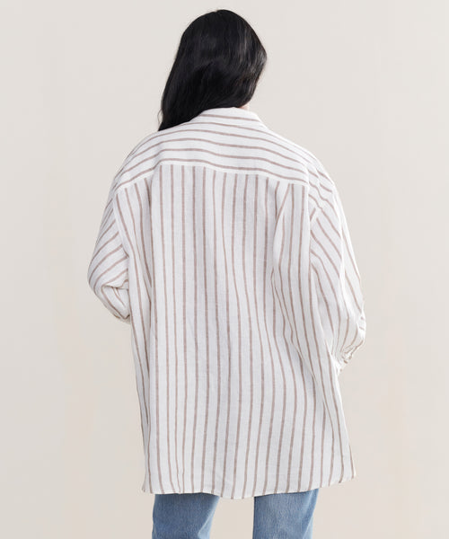 A person with long dark hair, seen from behind, wears the Linen Relaxed Oversized Shirt with vertical taupe stripes and light blue jeans, standing against a plain light background.