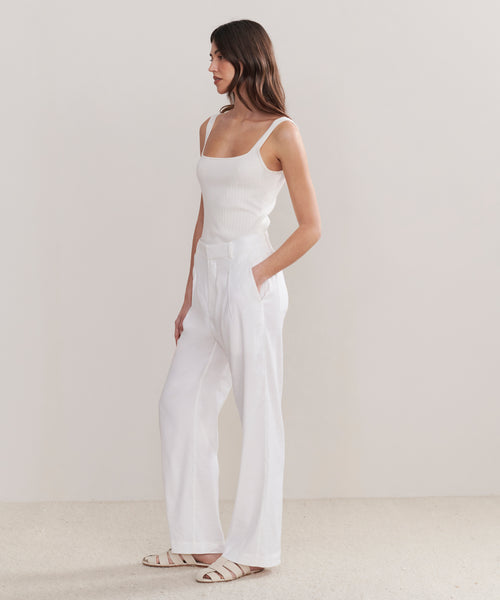 A woman with long brown hair faces left in a minimalist room, wearing a white sleeveless ribbed top, Riley Trouser—relaxed-fit, high-waisted linen trousers with pockets—and white flat sandals.