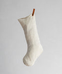 The Linen Stocking, made of ivory linen and featuring a brown leather hanging tab, is shown against a light gray background for a simple and elegant holiday accent.