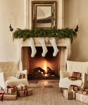 A cozy living room with a lit fireplace, three Linen Stockings with leather tabs hanging from a greenery-adorned mantel, two white armchairs, and wrapped gifts tied with red and white ribbons.