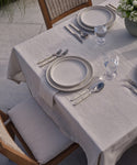 An outdoor table set with cream plates, silverware, and water glasses rests on a Linen Tablecloth with subtle black stripes. Wooden chairs with woven backs and cushions surround the setting, and a bowl of flowers decorates one end.