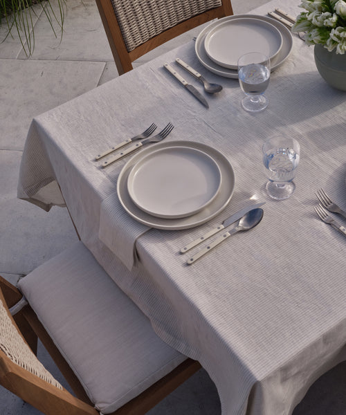 An outdoor table set with cream plates, silverware, and water glasses rests on a Linen Tablecloth with subtle black stripes. Wooden chairs with woven backs and cushions surround the setting, and a bowl of flowers decorates one end.