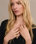 A woman with long blonde hair in a black top looks to the side, her hands near her chest, showcasing the Liv Rose Cut Ring along with several gold bands. She has a calm and thoughtful expression.