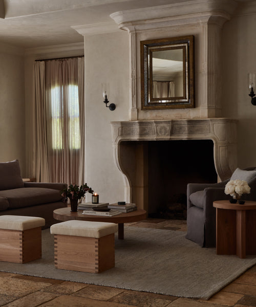 A cozy living room with a stone fireplace, large mirror, two armchairs, wooden stools, round coffee table, side table, neutral decor, and a Lodge Handwoven Rug beneath soft natural light from the window.