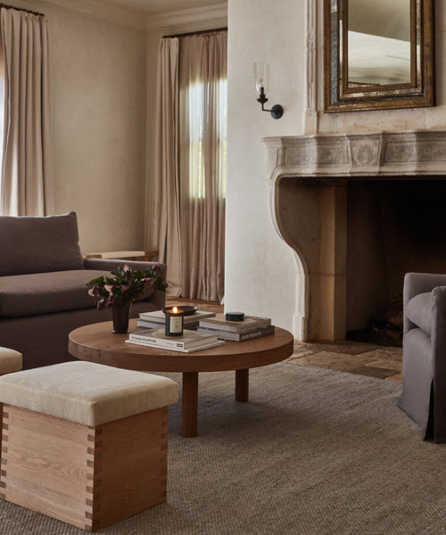 A cozy living room in neutral tones, featuring a stone fireplace, round wooden coffee table with books and a lit candle, two ottomans, gray sofa atop the Lodge Handwoven Rug, beige curtains, and a large mirror above the fireplace.
