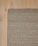 A close-up of the Lodge Handwoven Rug in light brown, featuring a textured pattern, is shown partially on a light wood floor. The left and bottom edges of the New Zealand wool rug align with the images edge.