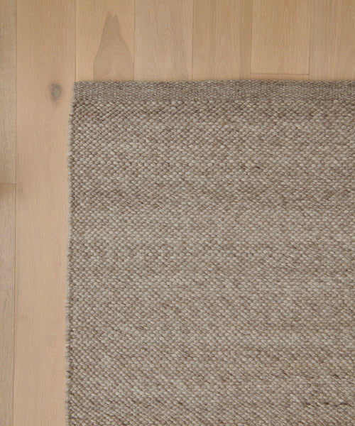 A close-up of the Lodge Handwoven Rug in light brown, featuring a textured pattern, is shown partially on a light wood floor. The left and bottom edges of the New Zealand wool rug align with the images edge.