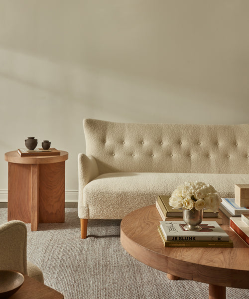 A beige tufted sofa sits next to a wooden side table with ceramic cups, while a round coffee table in the foreground holds stacked books and a vase of white flowers atop the Lodge Handwoven Rug, complementing the minimalist, neutral decor.