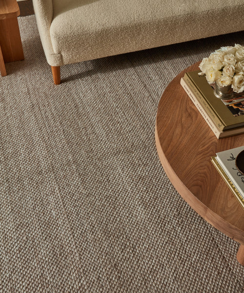 A cozy living room corner features a beige textured sofa, a round wooden coffee table with books and white roses, all set on the Lodge Handwoven Rug in light brown tones.