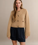 A woman wearing a beige button-up cardigan in lightweight fabric with front pockets and the Logan Short stands against a plain, light-colored background.
