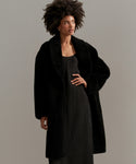 A woman with natural curly hair wears the Long Shearling Yves Shawl Coat over a black dress, standing against a plain, light gray background.