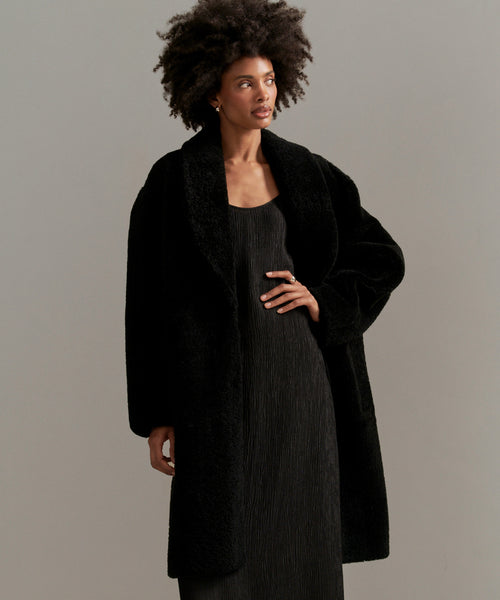A woman with natural curly hair wears the Long Shearling Yves Shawl Coat over a black dress, standing against a plain, light gray background.