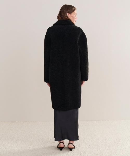 A woman stands facing away, wearing the Long Shearling Yves Shawl Coat over a black ankle-length skirt and black heeled sandals, against a plain light-colored background.