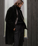 A person with shoulder-length brown hair stands outside a dark wooden building, wearing a Long Shearling Yves Shawl Coat over a black turtleneck and trousers, looking down with a neutral expression.