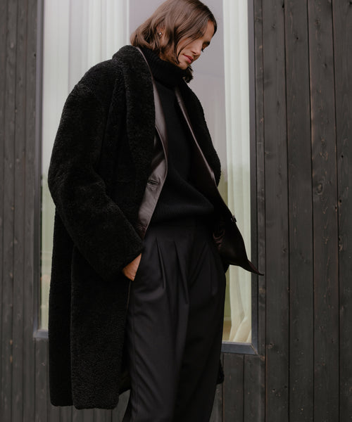 A person with shoulder-length brown hair stands outside a dark wooden building, wearing a Long Shearling Yves Shawl Coat over a black turtleneck and trousers, looking down with a neutral expression.