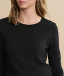 A woman with wavy, shoulder-length blonde hair stands against a beige background wearing the Long-Sleeve Slim Tee—a black, 100% cotton slim-fit top. Her face is partially visible with a neutral expression.