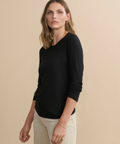 A woman with wavy, shoulder-length blonde hair wears the Long-Sleeve Slim Tee in black paired with light beige pants. She stands against a plain beige background, gazing confidently at the camera.