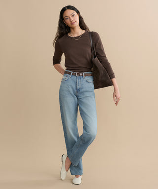 A woman with long dark hair wears the Long-Sleeve Slim Tee in brown, paired with light blue high-waisted jeans, white shoes, and a brown shoulder bag. She stands relaxed against a plain beige background.