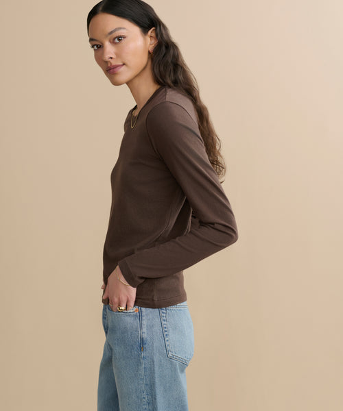 A woman with long dark hair models the Long-Sleeve Slim Tee and light blue jeans, standing sideways against a beige background and looking at the camera with a neutral expression.