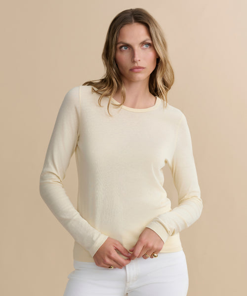 A woman with wavy light brown hair wears the Long-Sleeve Slim Tee and white pants, standing against a beige background with a neutral expression.