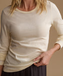 A close-up of a woman wearing a cream-colored long-sleeve crewneck top, holding the hem to show the fabric drape. The background is a solid warm beige studio backdrop.