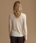 A woman viewed from behind wearing a light cream long-sleeve knit top and dark pants, standing against a solid warm beige studio background.
