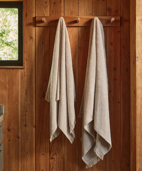 Two Loom Bath Towels hang side by side on wooden hooks against a wooden wall. Natural light from a small window enhances the cozy, rustic feel—perfect for these super-soft towels made in Portugal.