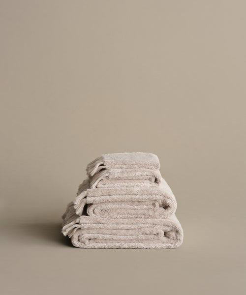 Three neatly folded Loom Bath Towels, expertly crafted in Portugal for exceptional softness, are stacked against a plain beige background—adding a touch of elegance to any bathroom.