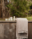 The Loom Bath Towel, crafted in Portugal with fringed edges, is displayed alongside skincare products on a rustic wooden fence, set against a lush forest backdrop.
