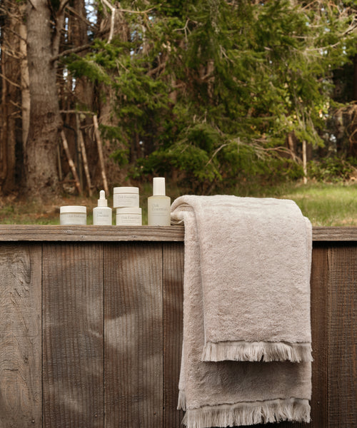 The Loom Bath Towel, crafted in Portugal with fringed edges, is displayed alongside skincare products on a rustic wooden fence, set against a lush forest backdrop.