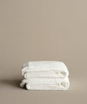 Two neatly folded Loom Bath Towels, crafted in Portugal, are stacked against a plain beige background, highlighting their luxurious softness and quality.