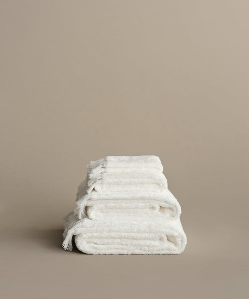 A neatly stacked pile of four white Loom Bath Towels with super-soft texture and fringed edges sits on a neutral beige surface against a matching beige background.