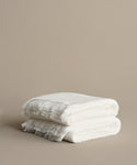 Two neatly folded Loom Bath Towels in super-soft white with fringed edges are stacked on a light beige surface, set against a plain beige background.