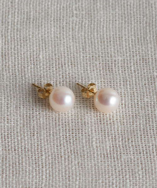 The Large Loren Pearl Studs are round freshwater pearl earrings with gold posts and backings, shown on a textured cream-colored fabric.