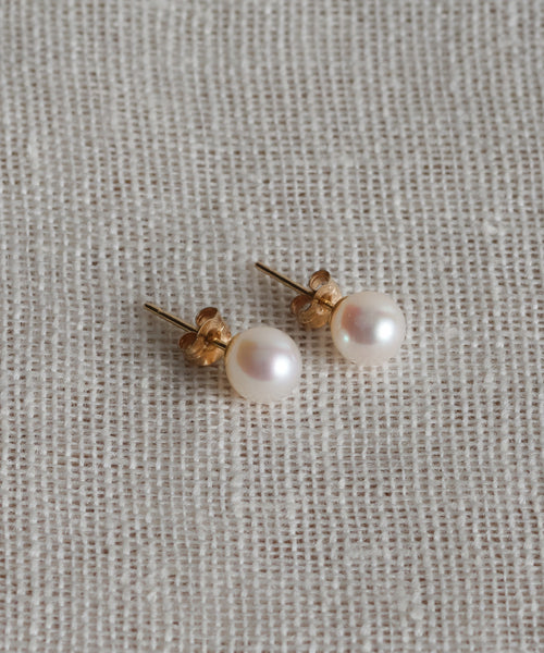 The Medium Loren Pearl Studs, crafted in 14k gold and featuring round freshwater pearls, rest elegantly on textured beige fabric.