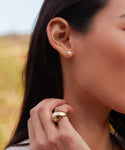 A womans ear is adorned with Medium Loren Pearl Studs and her hand near her neck displays a large, shiny 14k gold ring, set against a softly blurred background.