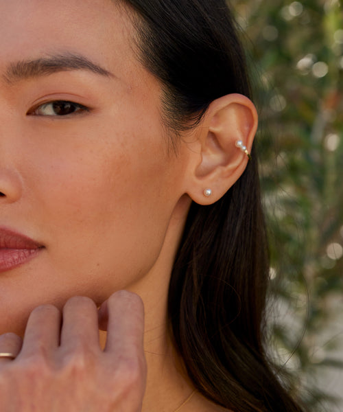 Close-up of a womans face and ear, highlighting smooth skin, minimal makeup, and gold jewelry: a lobe stud, two cartilage studs, and the Loren Pearl Ear Cuff. Her dark hair is tucked back as her hand gently rests on her chin.
