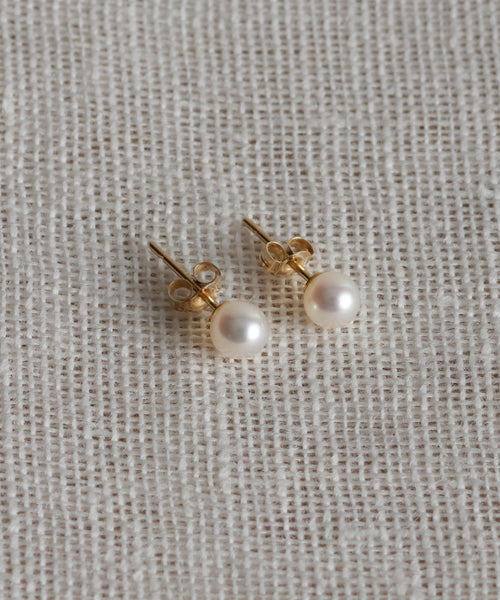 A pair of Small Loren Pearl Studs by Jenni Kayne Jewelry, featuring gold and round white pearls, rests on a textured off-white fabric background.