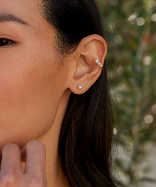 Close-up of a woman’s ear adorned with Jenni Kayne’s Small Loren Pearl Studs in her lobe and two small gold hoops in her upper cartilage. Her long dark hair frames her face as she rests her hand on her chin, with blurred green foliage behind.