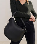 A person in a dark green sweater and black pants holds the large, black Loren Tote over their shoulder. The Italian-made bag stands out against a plain, light-colored background.