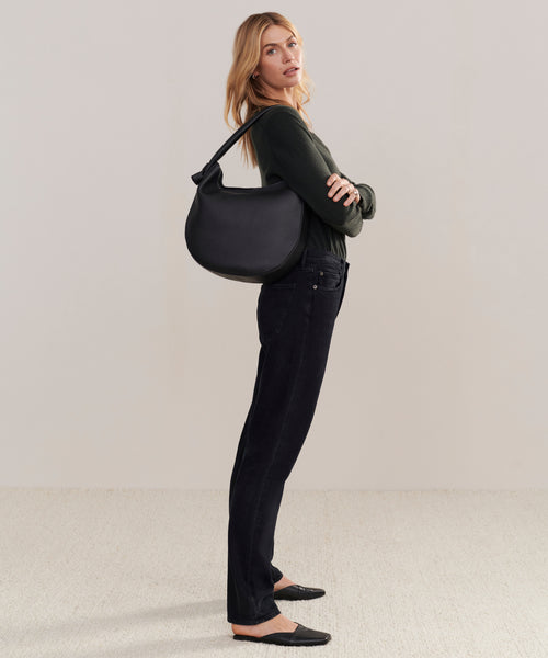 A woman stands sideways with arms crossed, wearing a dark green long-sleeve top, black jeans, and flats, carrying the Loren Tote—a leather tote made in Italy. The background is plain and neutral.
